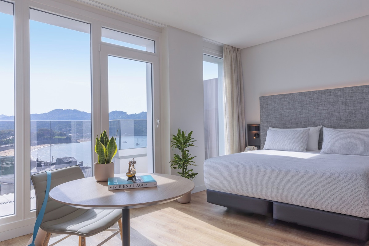 Une chambre moderne avec un grand lit, une table ronde avec une chaise, et une vue sur une étendue d’eau grâce à de grandes fenêtres.