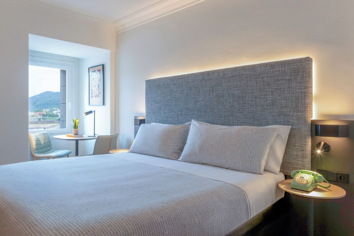 Ein Hotelzimmer mit einem großen Bett, einem Nachttisch mit Lampe, einem Sessel und Blick ins Freie.