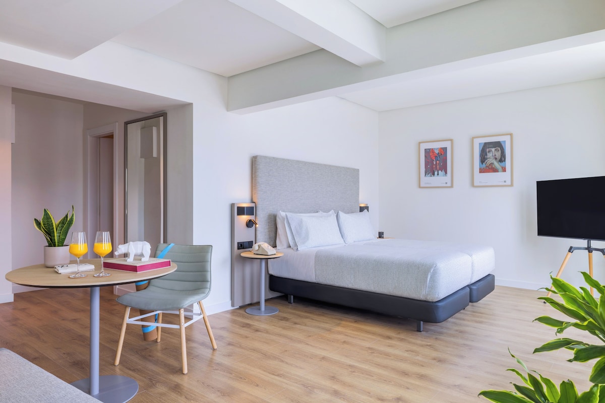 Une chambre d’hôtel moderne équipée d’un lit, d’une table de chevet, d’une chaise et d’une petite table sur laquelle se trouvent des boissons et un livre.