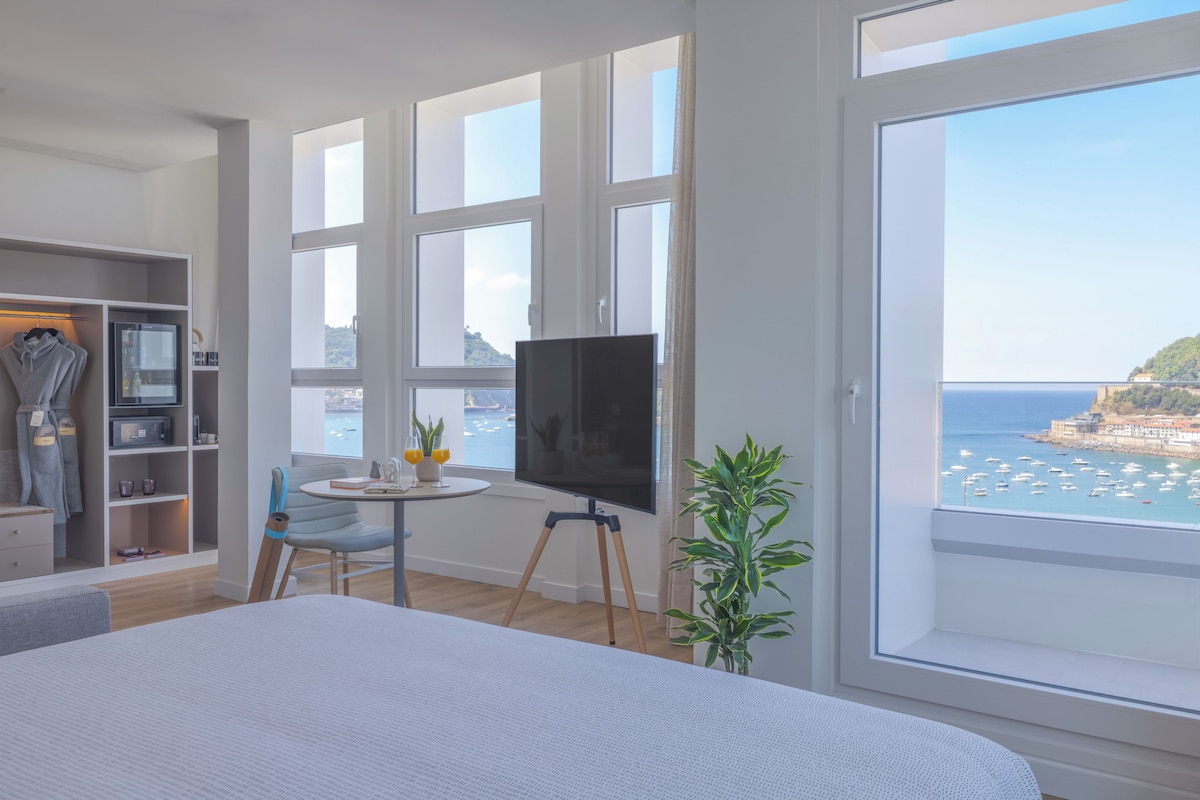 Une chambre dotée d’un grand lit, d’une télévision, d’un bureau avec une chaise, d’une armoire et offrant une vue sur la mer grâce à de grandes fenêtres.