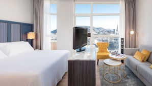A hotel room with a bed, a TV, a sofa, a small table, and a view of a cityscape through large windows.