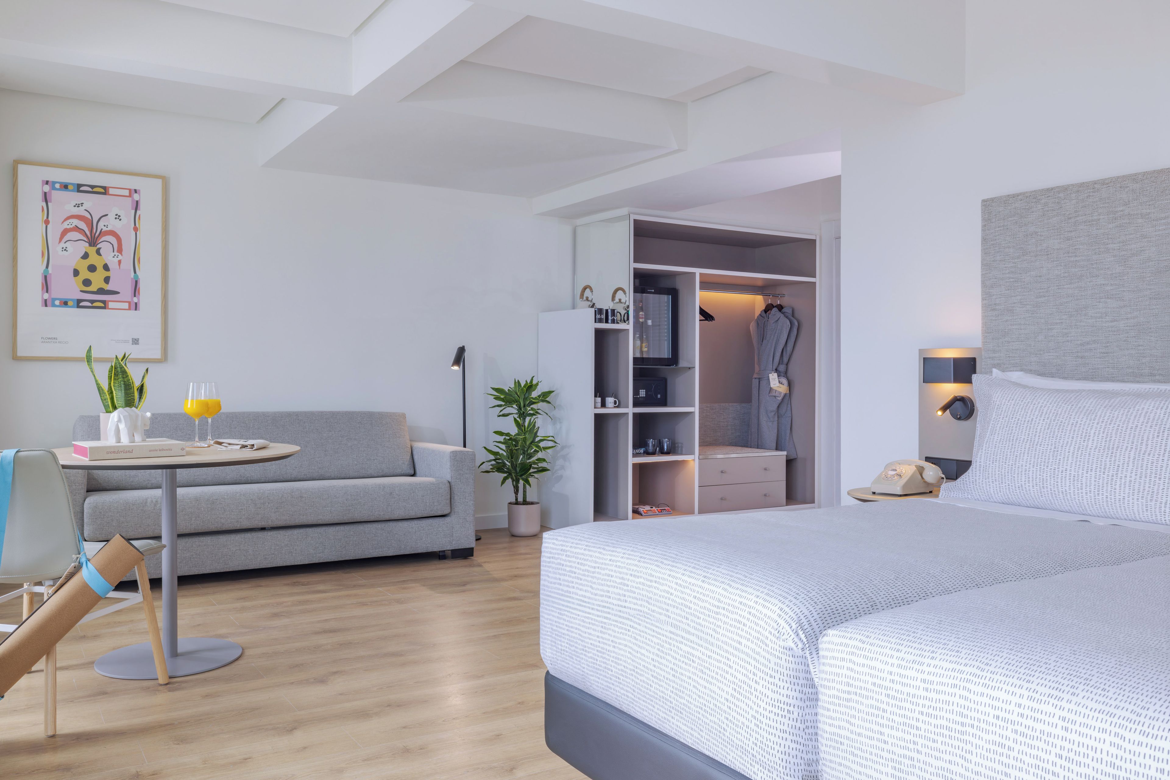 Una habitación de hotel moderna con una cama, un sofá, una pequeña mesa con una planta y un libro, un armario con ropa y un cuadro en la pared.