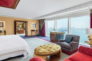 A hotel room with a large bed, a wooden ottoman, a red sofa, a patterned armchair, and a view of a city skyline through large windows.