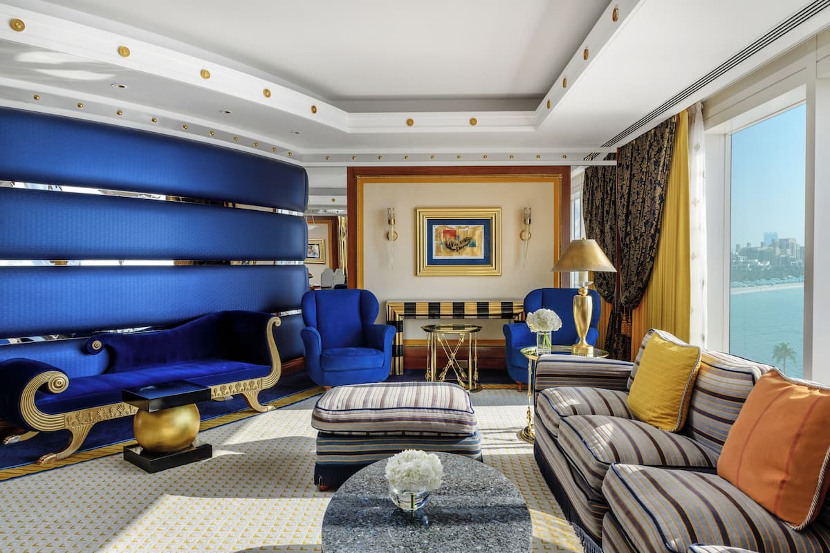 Ein luxuriöses Wohnzimmer mit einem blauen Sofa, einem gestreiften Sofa und einem gestreiften Sessel. Ein runder Couchtisch mit einer Blumenvase steht im Raum. An der Wand hängt ein Gemälde, und auf einem Beistelltisch steht eine Lampe.