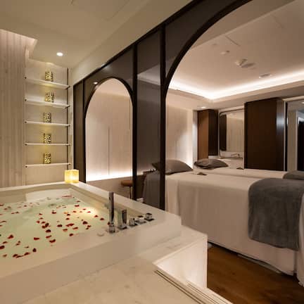 A modern spa room with a large bathtub, two beds, and a wooden panel wall.