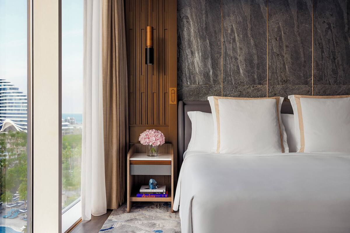 Una habitación de hotel moderna con una cama grande, una mesita de noche con un florero y vistas a un paisaje urbano a través de amplios ventanales.