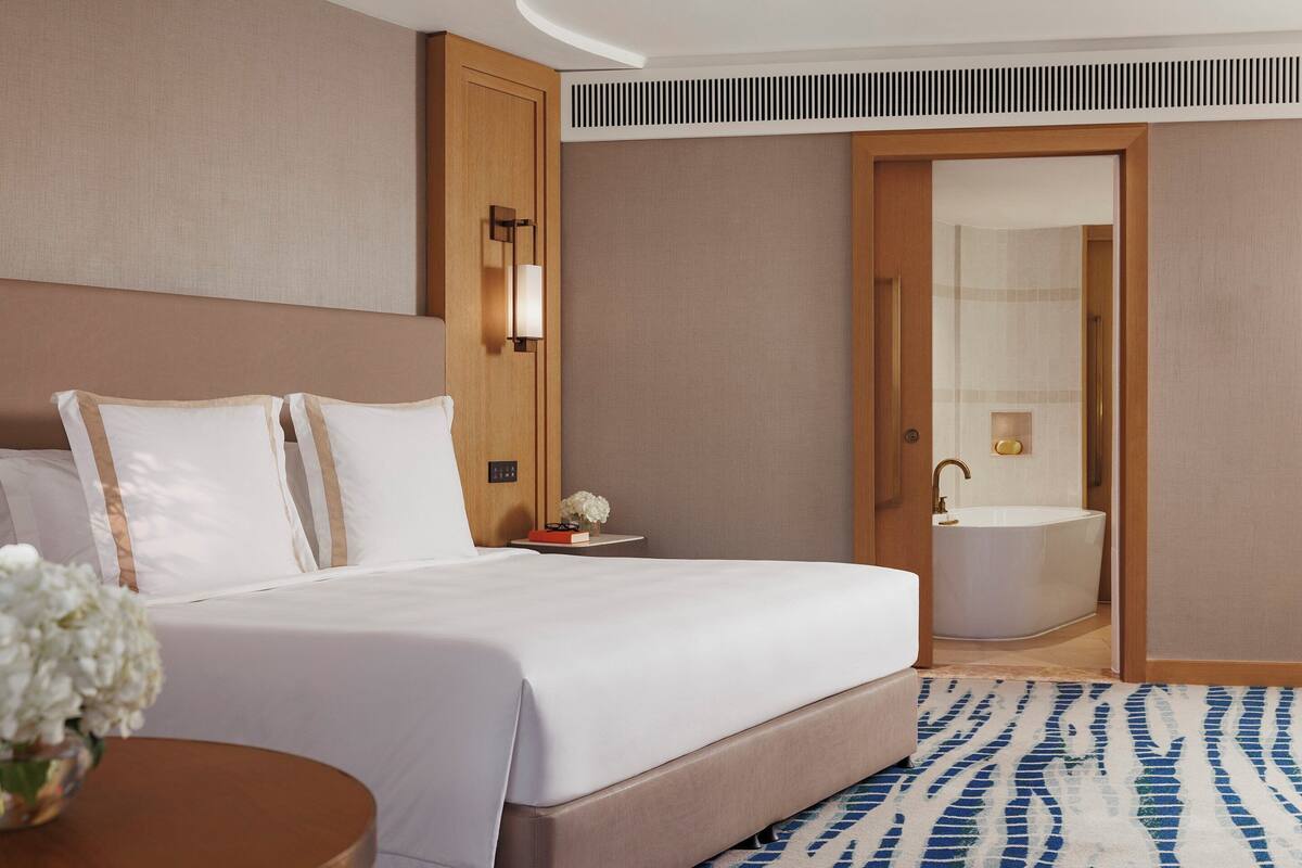 Habitación de hotel con una cama grande, una mesita de noche con un florero y un baño con bañera, además de una alfombra estampada.