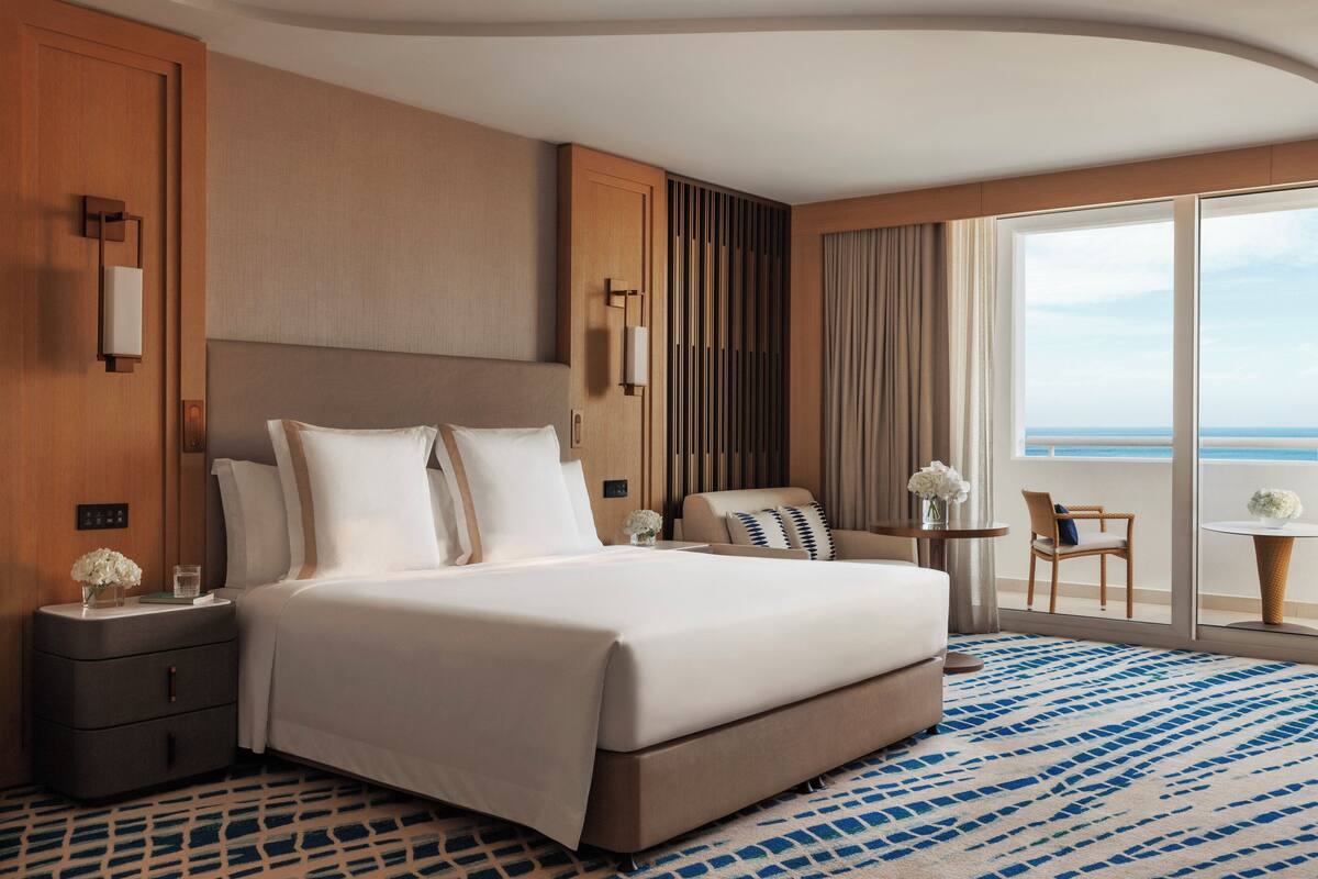 Habitación de hotel con una cama grande, una mesita de noche, una silla, una pequeña mesa y vista al océano.