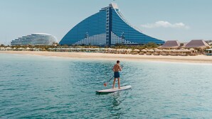 En mand står på paddleboard på et roligt hav med en moderne bygning i baggrunden.
