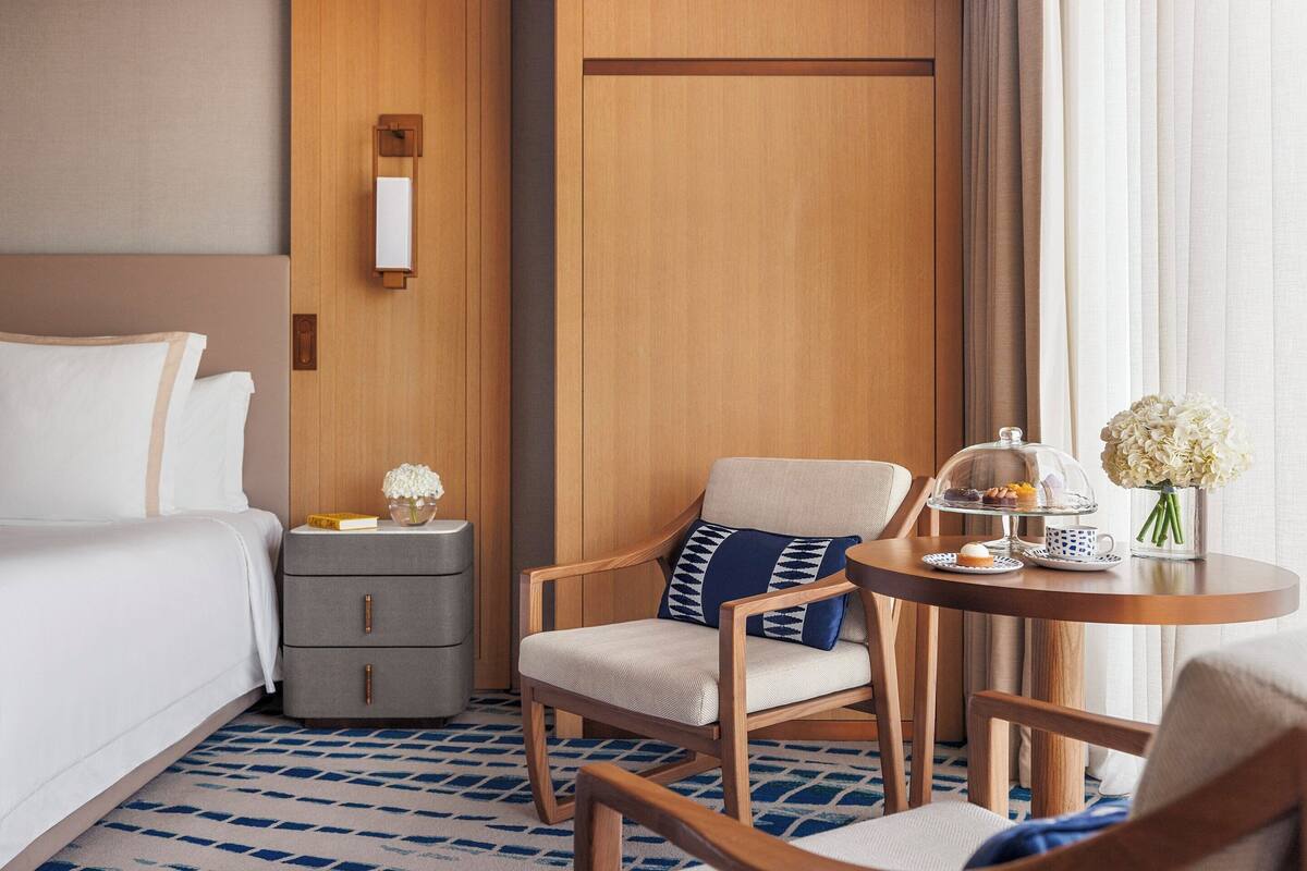 Habitación de hotel con cama, mesita de noche, una silla, una mesa con tetera y flores, y una puerta de madera.