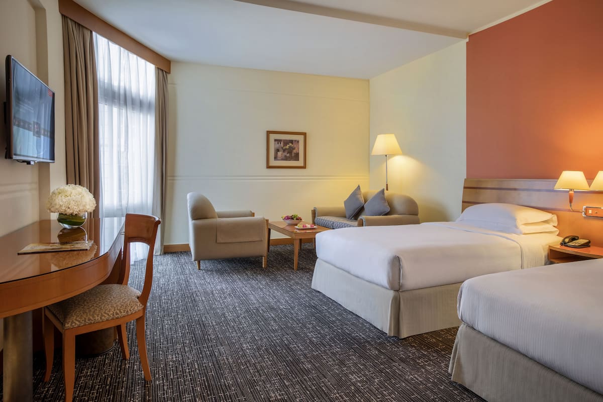 A hotel room with two beds, a desk, a chair, a TV, and a lamp.