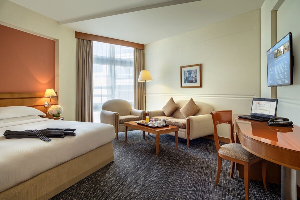 A hotel room with a bed, two armchairs, a sofa, a coffee table, a desk with a laptop, a lamp, and a TV on the wall.