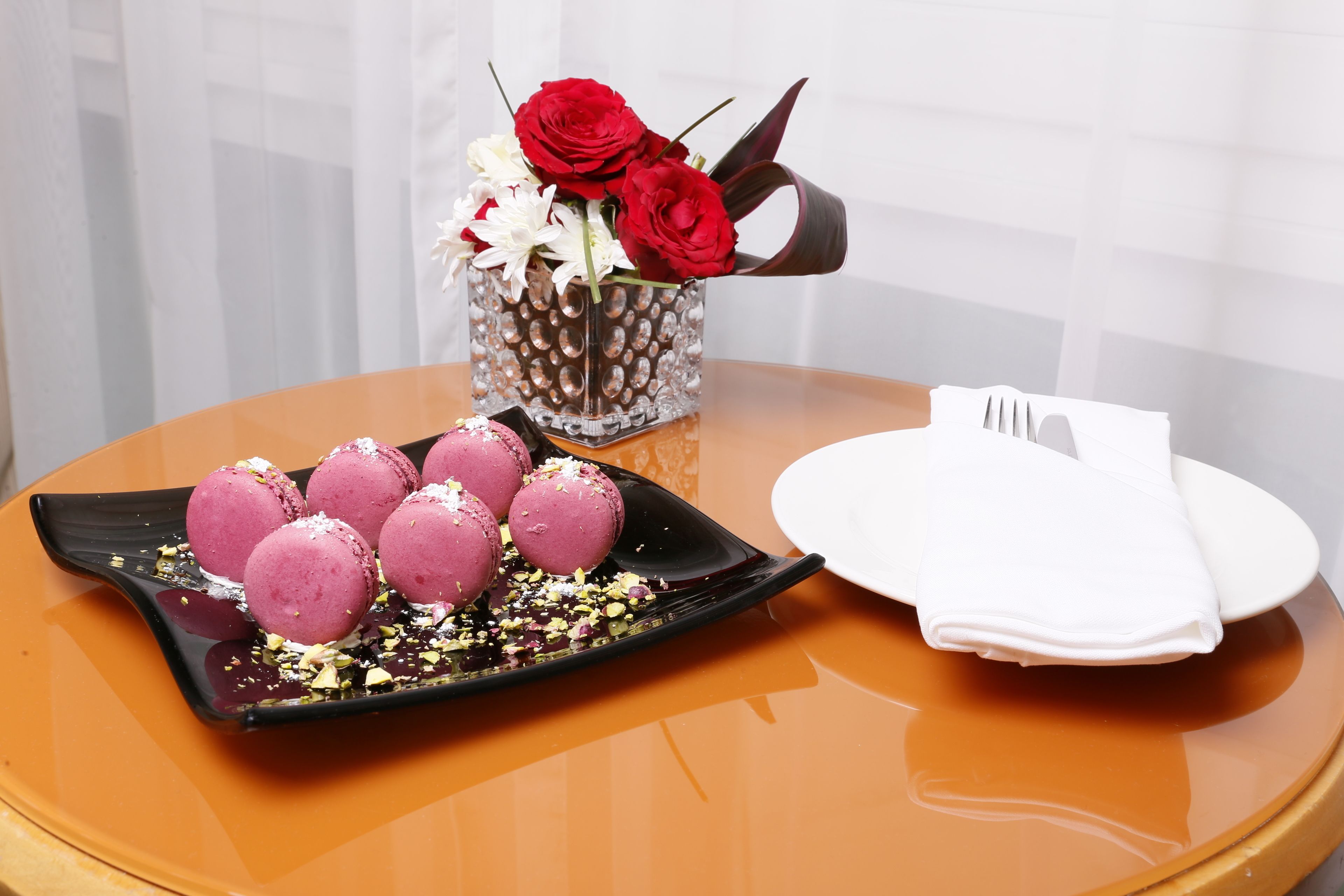 Una mesa con una bandeja de macarons rosas, una jarra de cristal con rosas rojas y una servilleta blanca doblada.