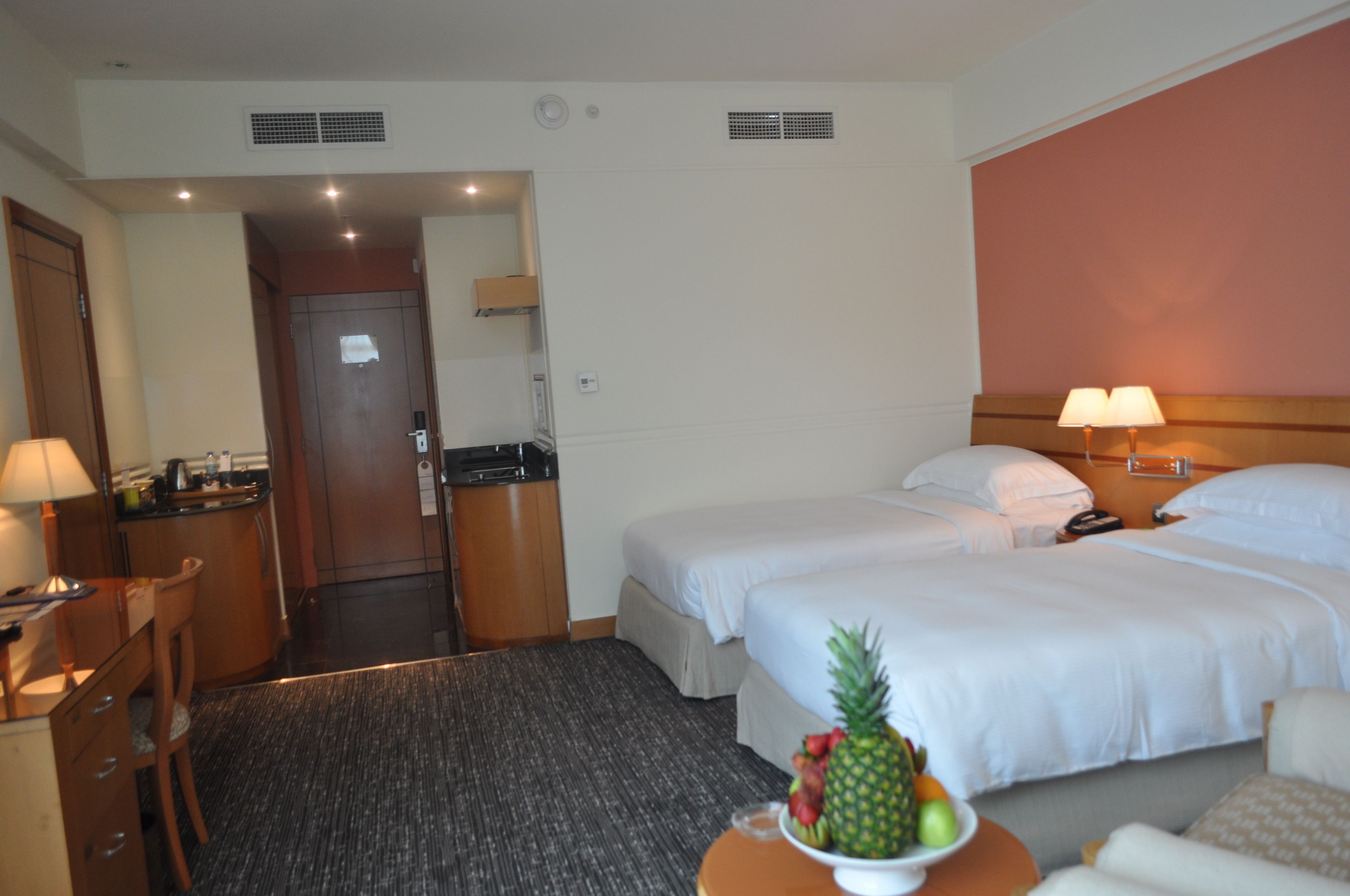 A hotel room with two beds, a desk with a chair, a small table with fruit, and a bathroom visible through an open door.