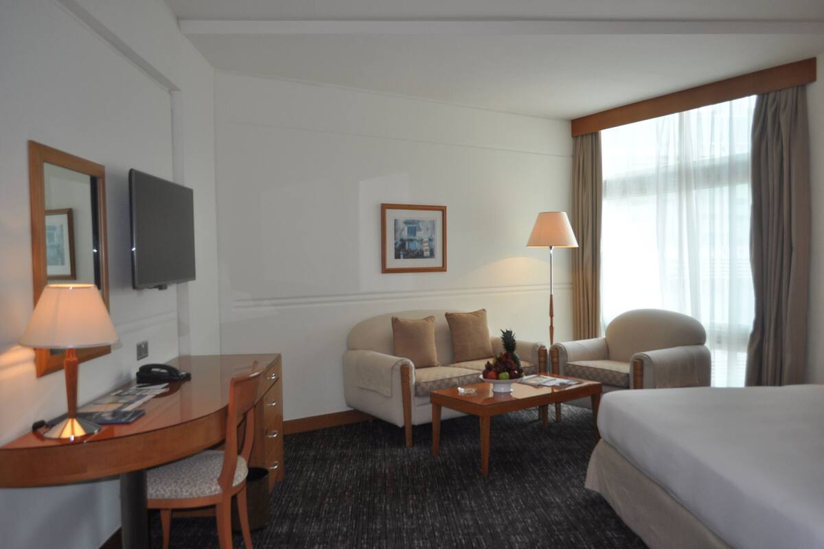 A hotel room with a bed, a desk with a lamp, a chair, a sofa, a coffee table, and a television.