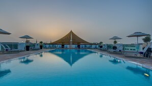 A swimming pool with lounge chairs and umbrellas, surrounded by buildings and a large tent structure. - J5 Hotels - Port Saeed (Dubai)