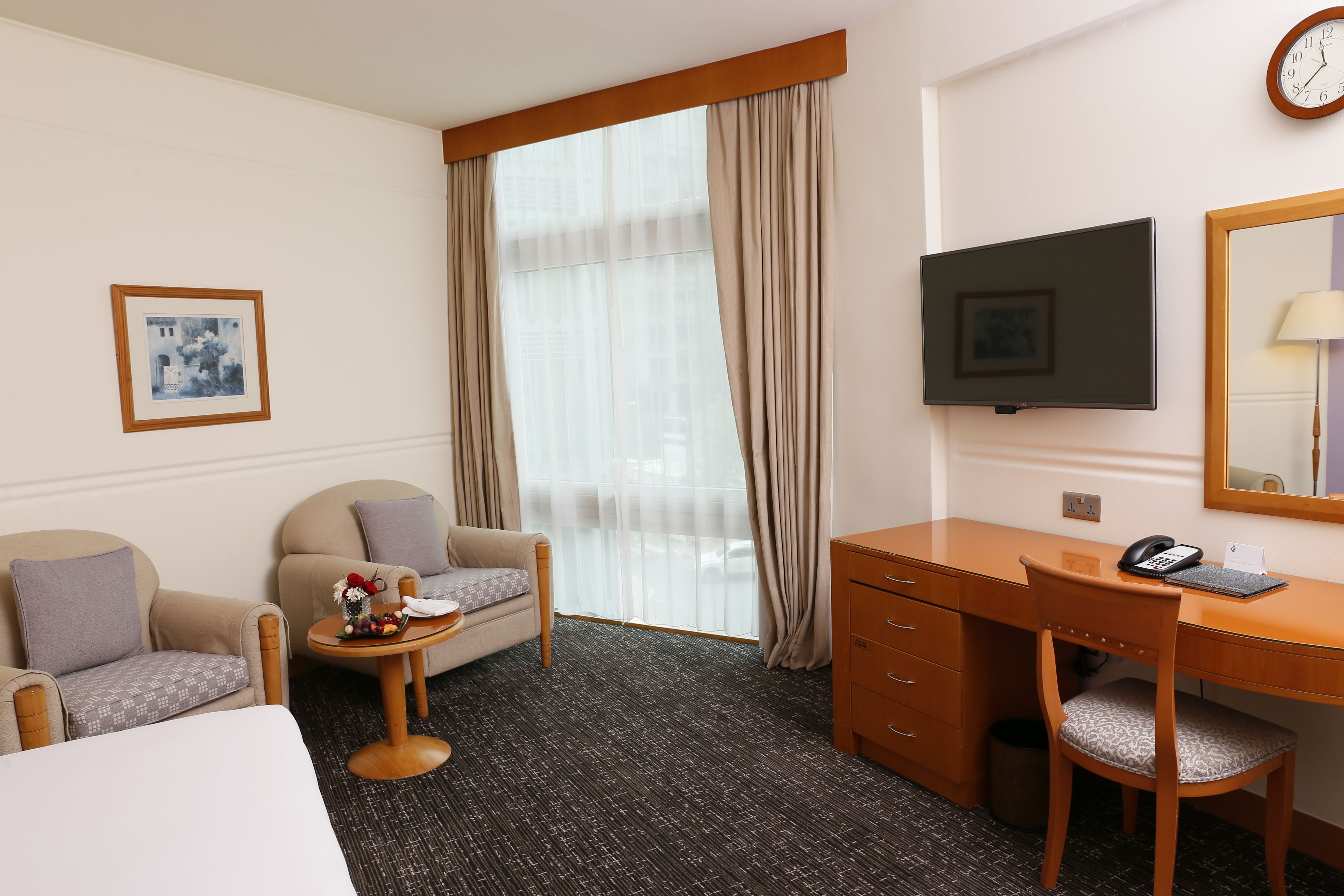 Een hotelkamer met een bed, twee fauteuils, een tafeltje met een bloevas, een bureau met een stoel, een televisie en een groot raam met gordijnen.