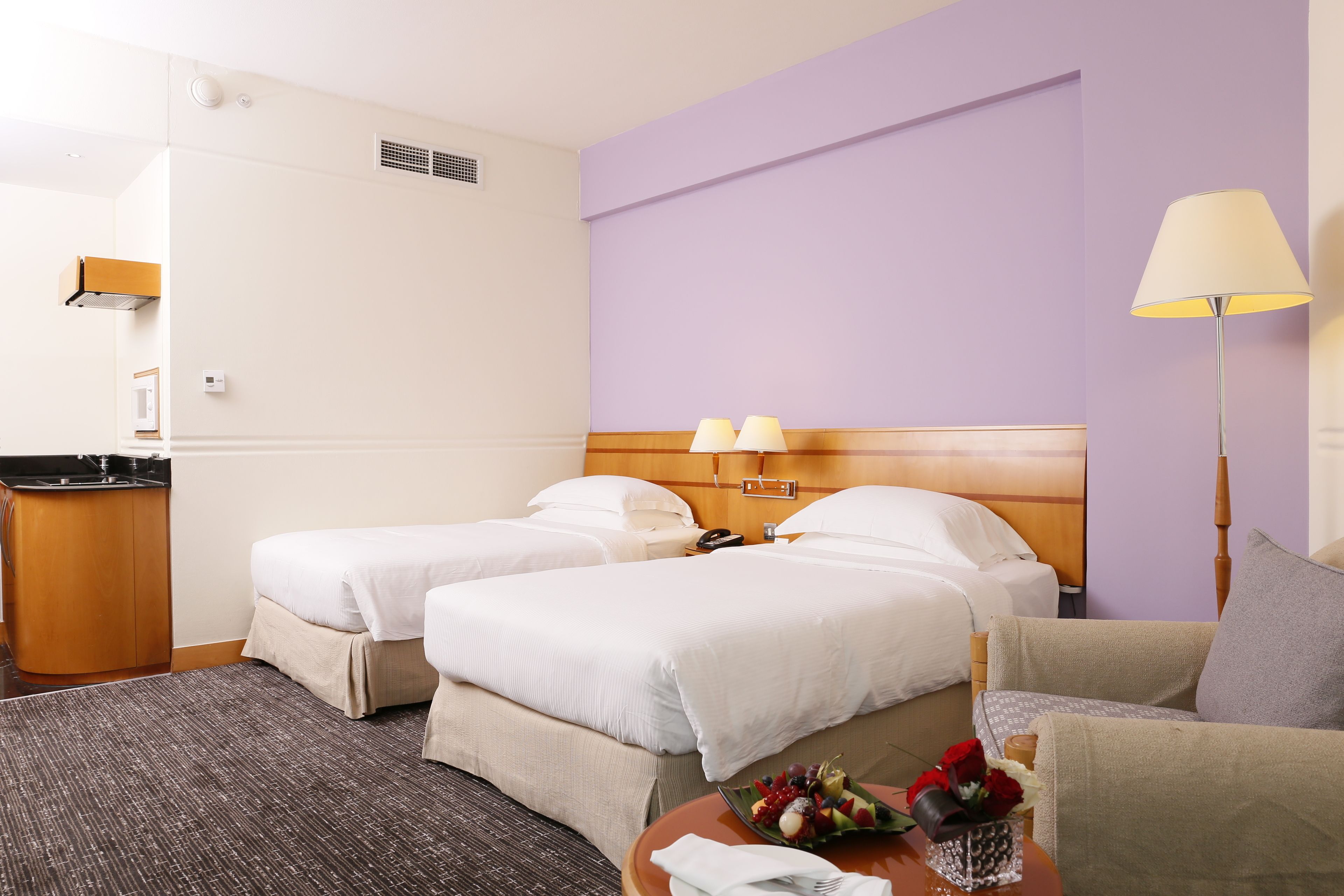 Hotellihuone, jossa on kaksi sänkyä, työpöytä, tuoli, lamppu ja hedelmäkori.