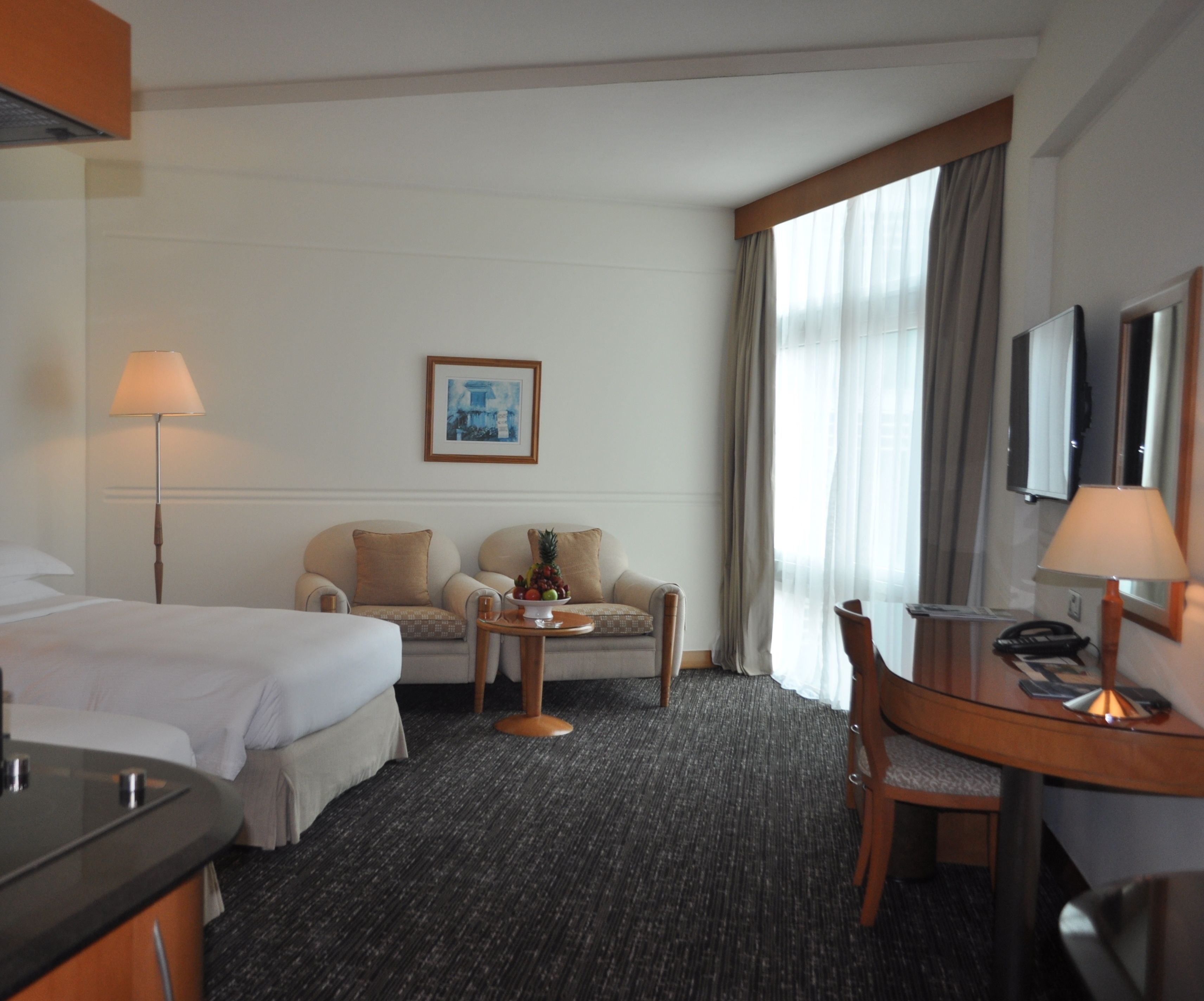 A hotel room with a bed, two armchairs, a small table with a fruit bowl, a desk with a lamp, and a large window with curtains.