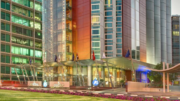 A modern hotel with a glass facade, multiple floors, and a prominent entrance with flags. - J5 Hotels - Port Saeed (Dubai)