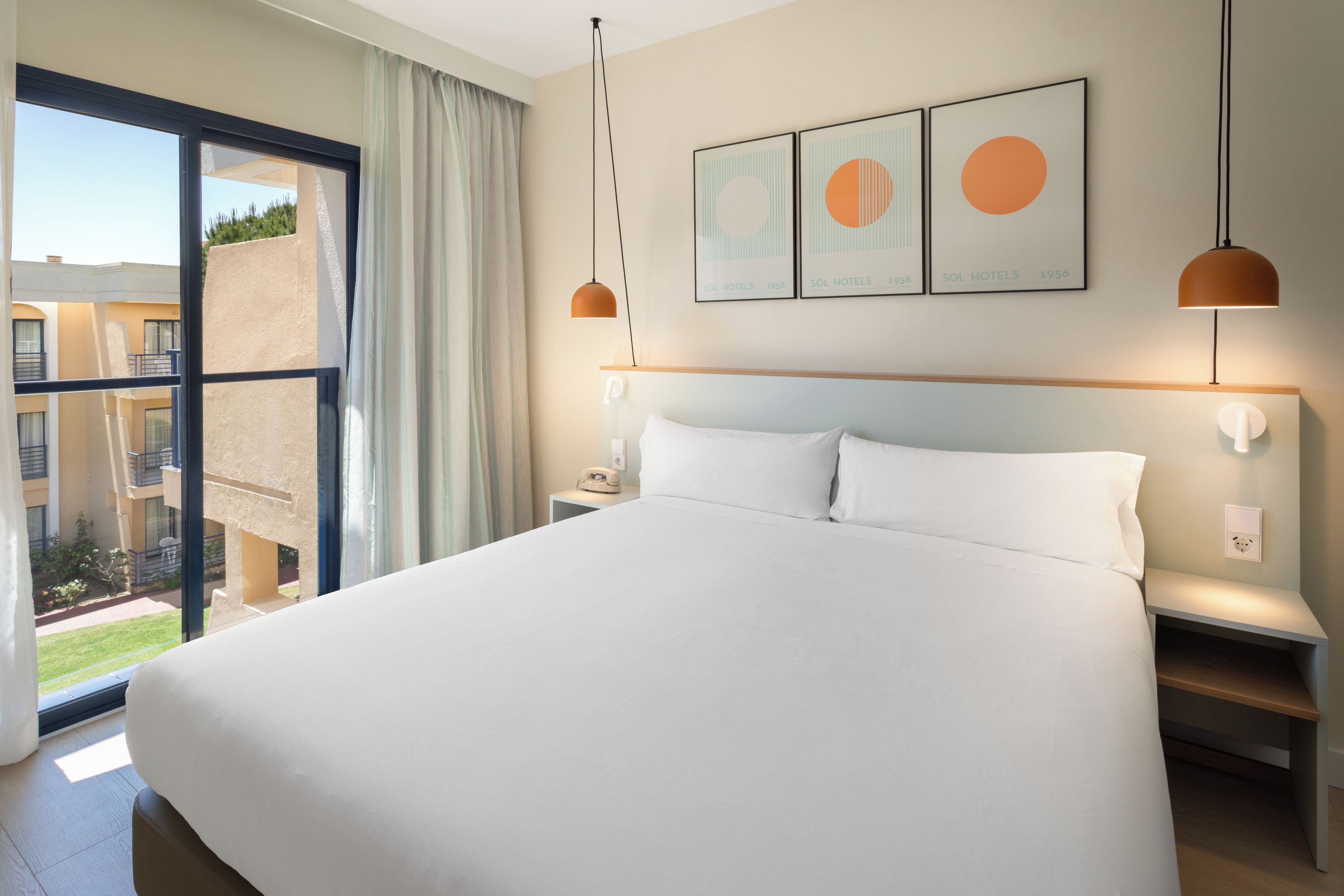 A hotel room with a large bed, a nightstand, a bedside table, a window with a view of the outside, and three framed pictures on the wall.