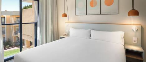 A hotel room with a large bed, a nightstand, a bedside table, a window with a view of the outside, and three framed pictures on the wall.