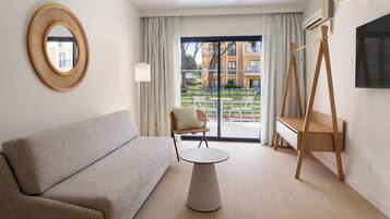 A modern hotel room with a sofa, a small round table, a chair, a TV, and a balcony with a view of buildings and greenery.