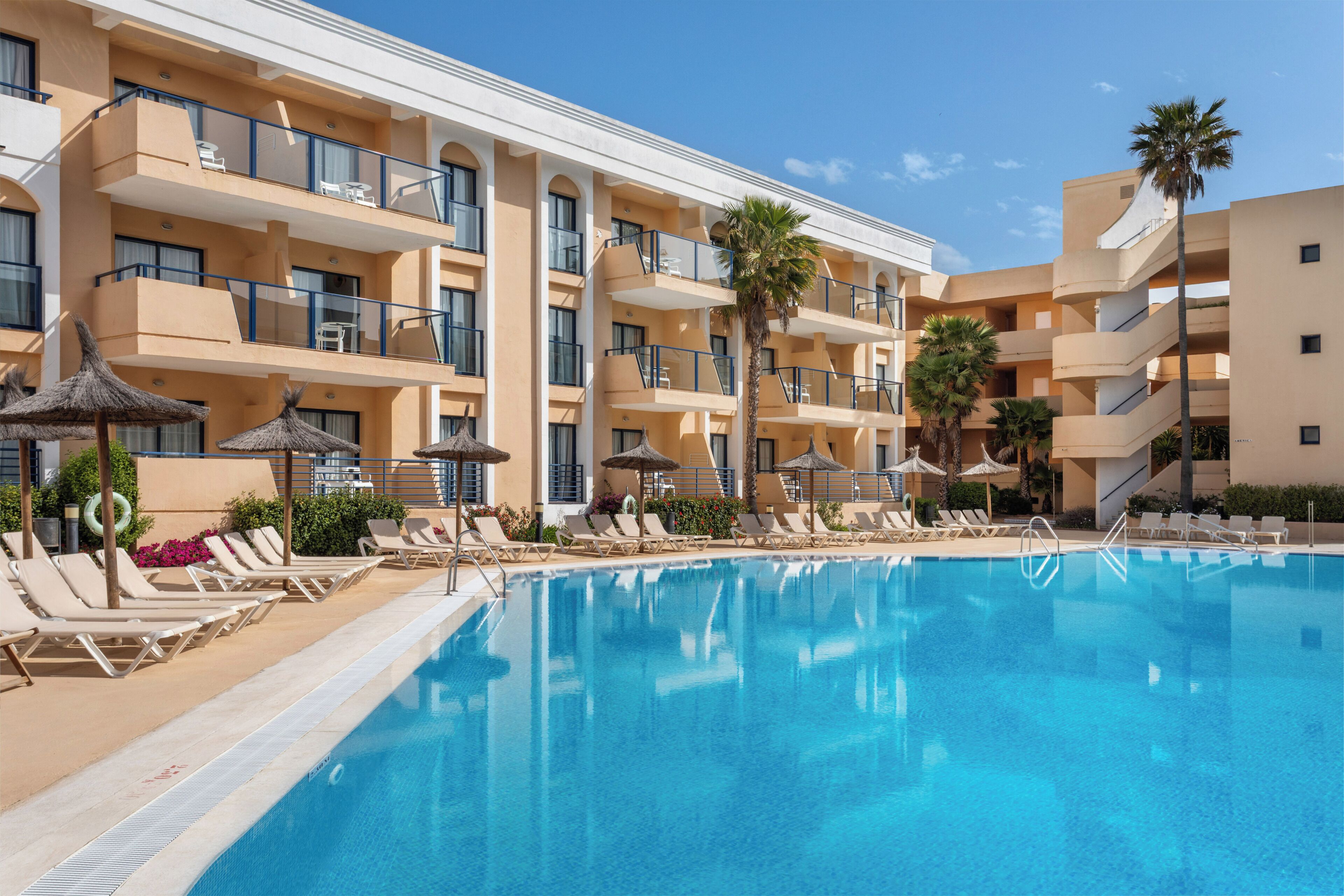 A hotel with a swimming pool, sun loungers, and palm trees.