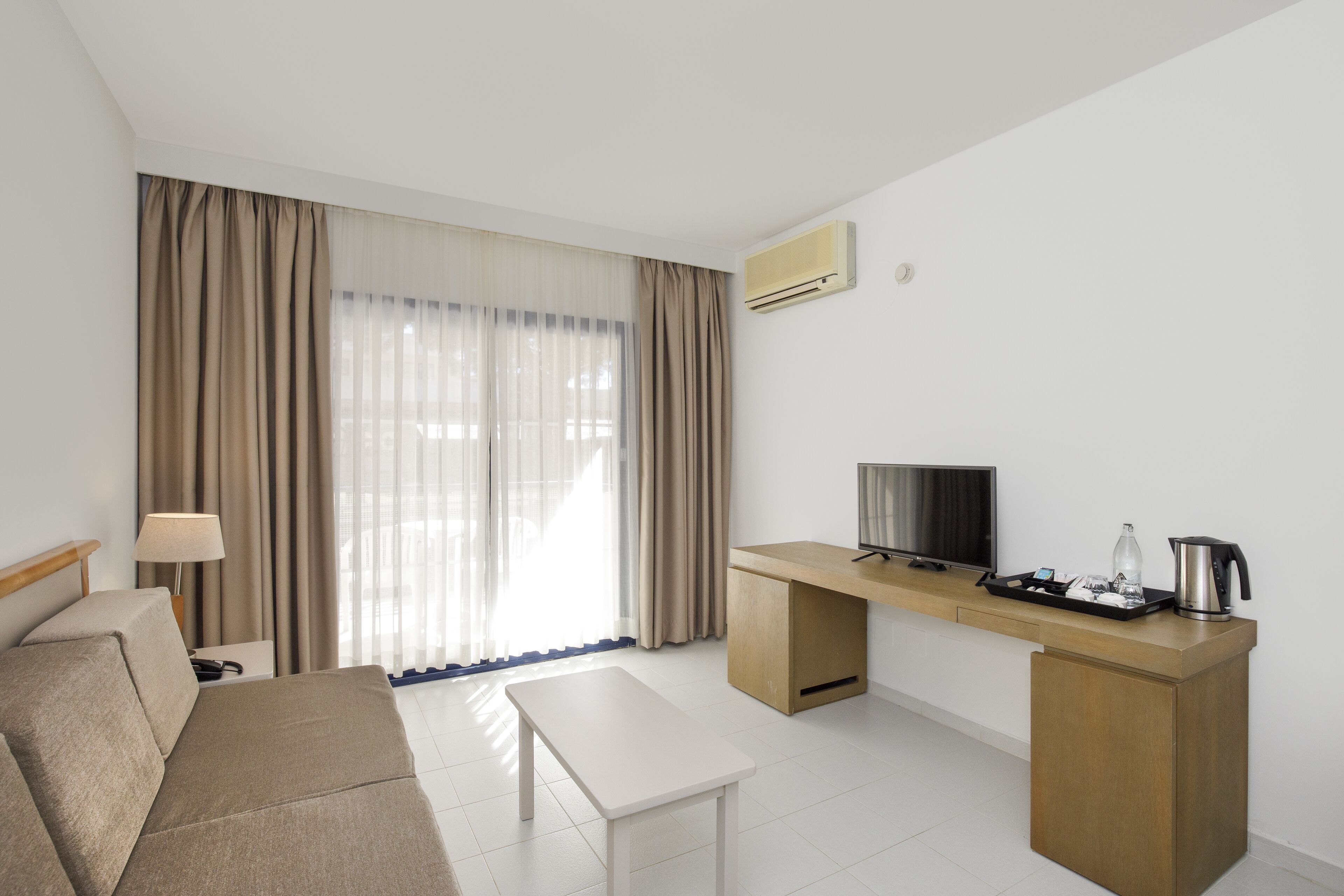 A modern hotel room with a sofa, a small table, a desk with a TV, and a coffee maker.