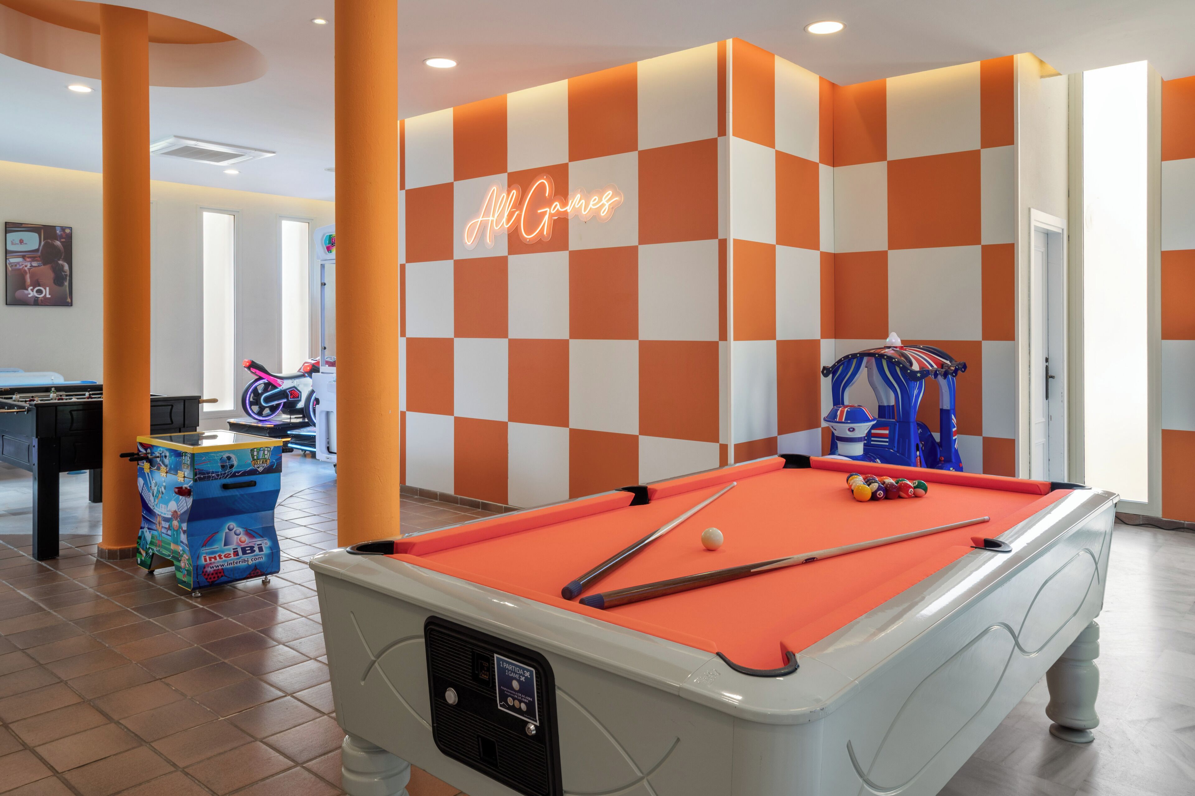 Una mesa de billar con un letrero de neón, una mesa de fútbol de mesa y una máquina de garra en una habitación muy iluminada con paredes a cuadros naranja y blanco.