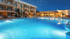 Zona de piscina de hotel con sillones de descanso, una piscina y un edificio con varios balcones.