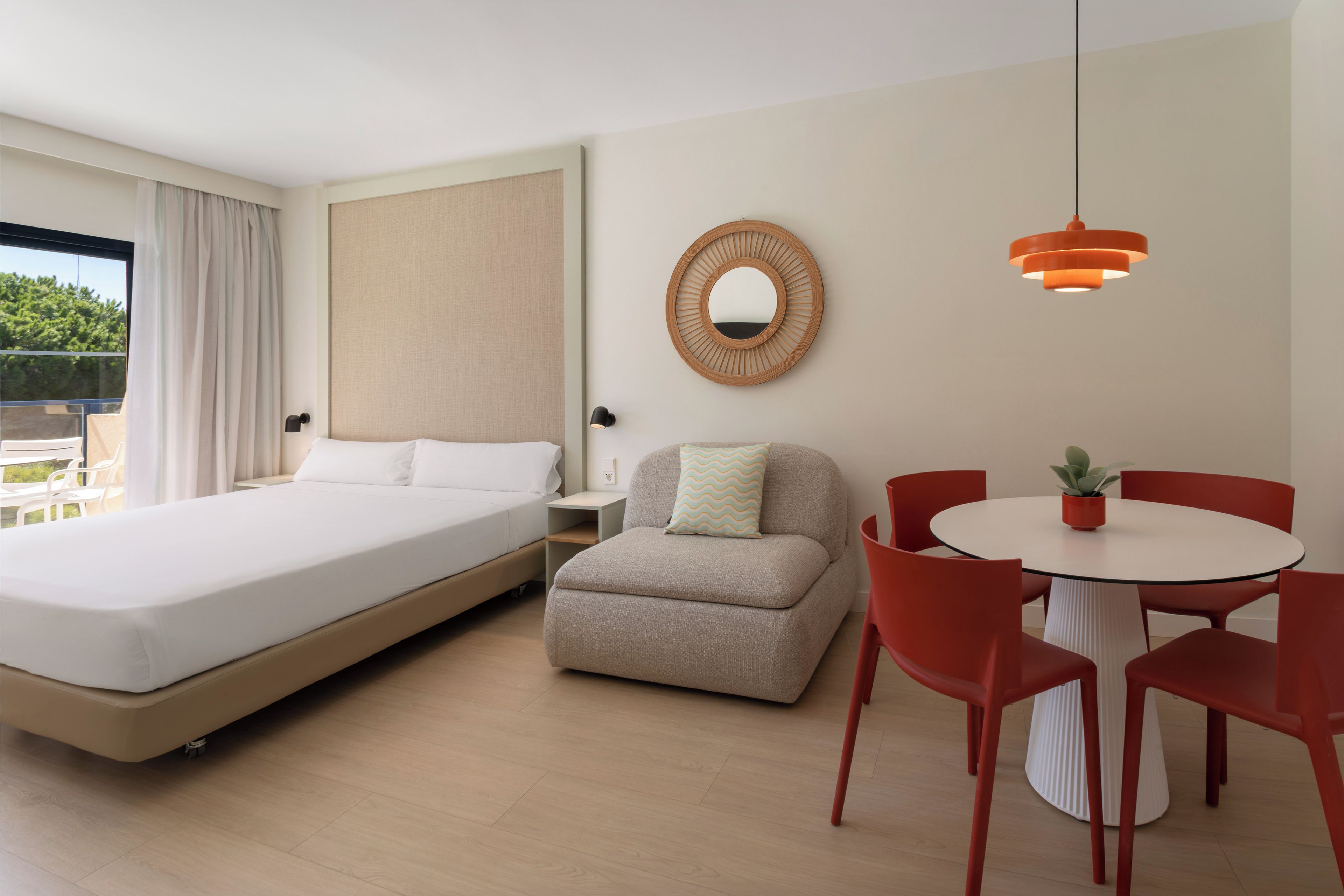 A hotel room with a bed, a sofa, a dining table with chairs, a mirror, and a hanging light fixture.