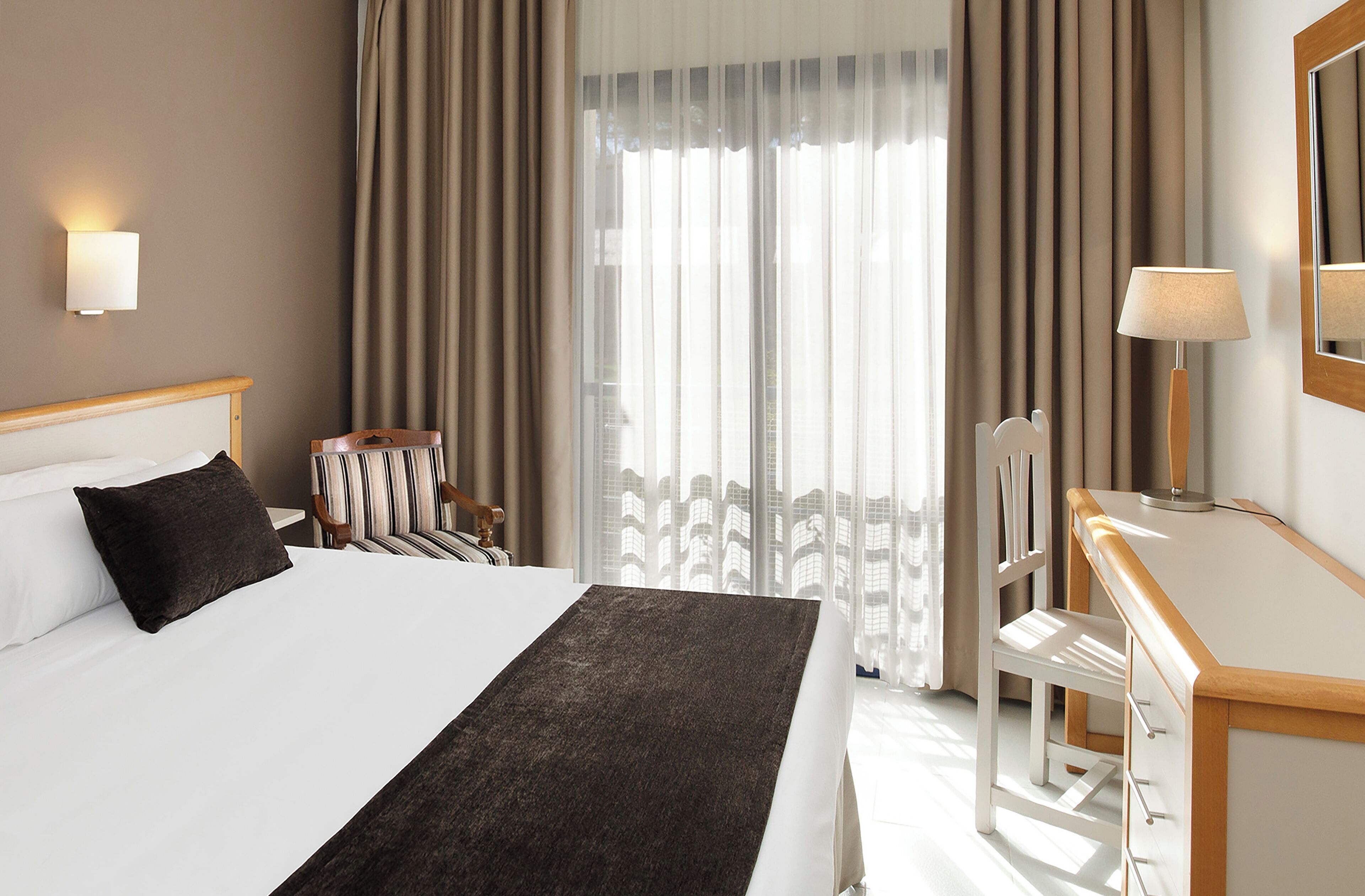 A hotel room with a bed, a desk, a chair, a lamp, and a view of buildings through the window.
