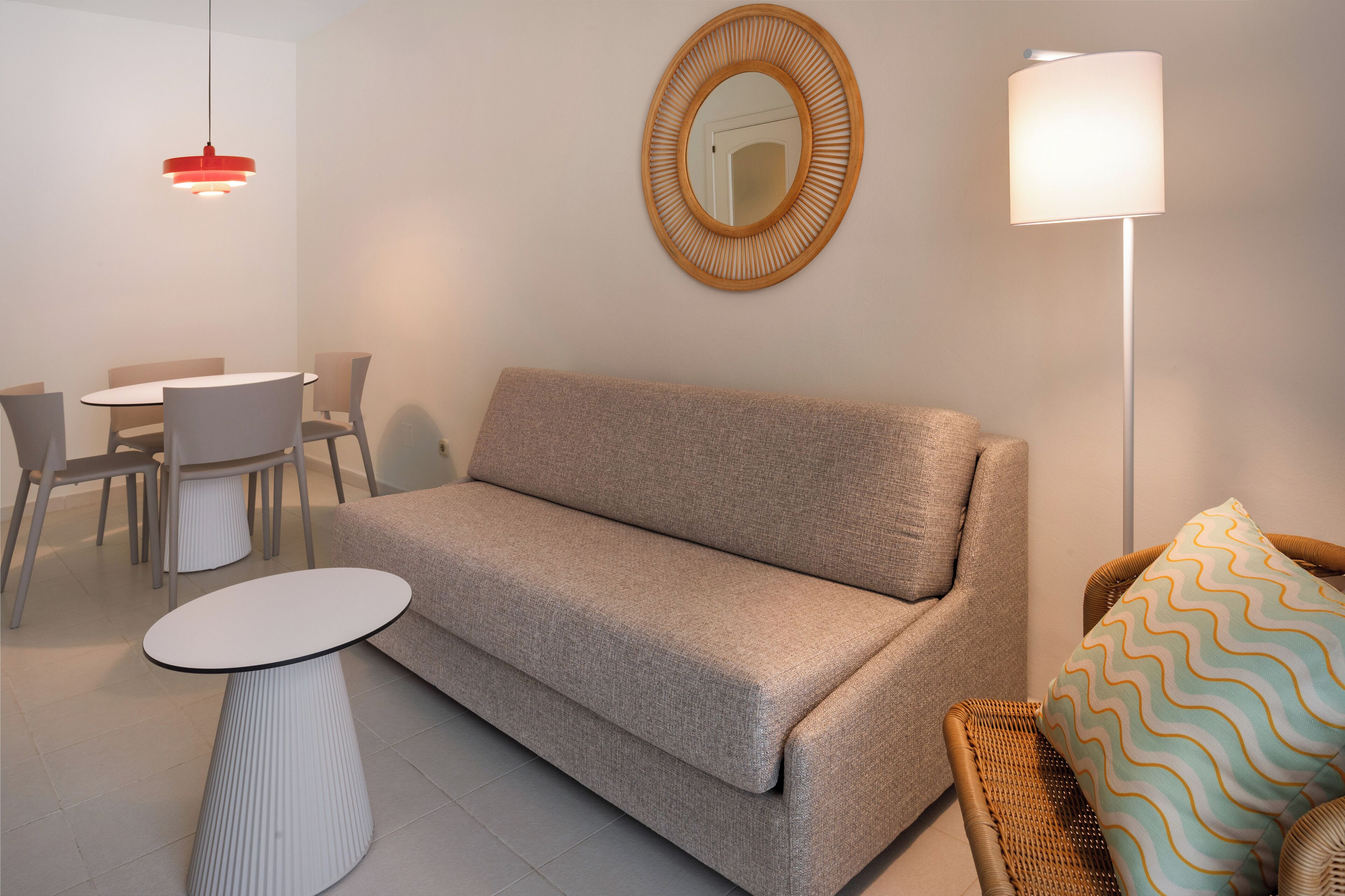 Uma sala de estar moderna com um sofá bege, um espelho redondo, um abajur, uma mesa de jantar com cadeiras e uma pequena mesa branca.