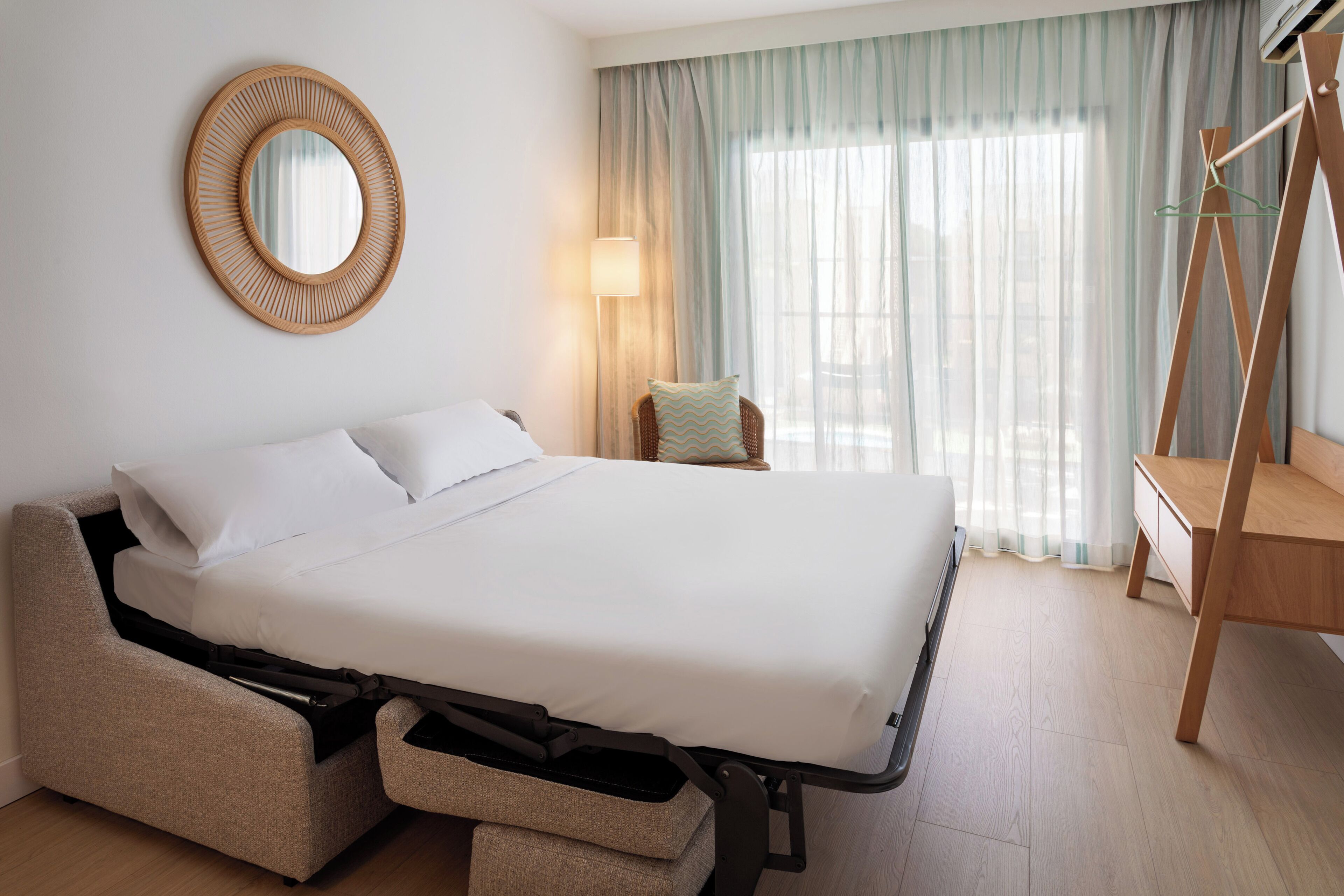 A modern hotel room with a convertible bed, a round mirror, a bedside lamp, a wooden desk, and a large window with curtains.