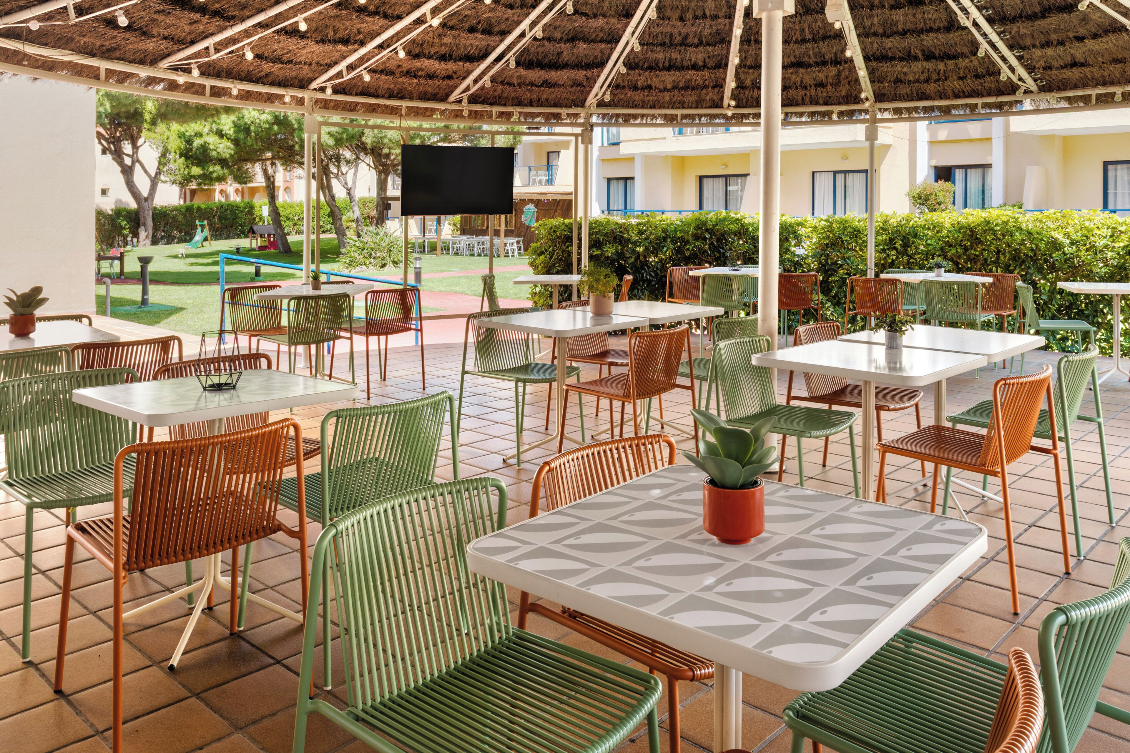 An outdoor patio area with tables and chairs, a thatched roof, and a view of a playground and building in the background.