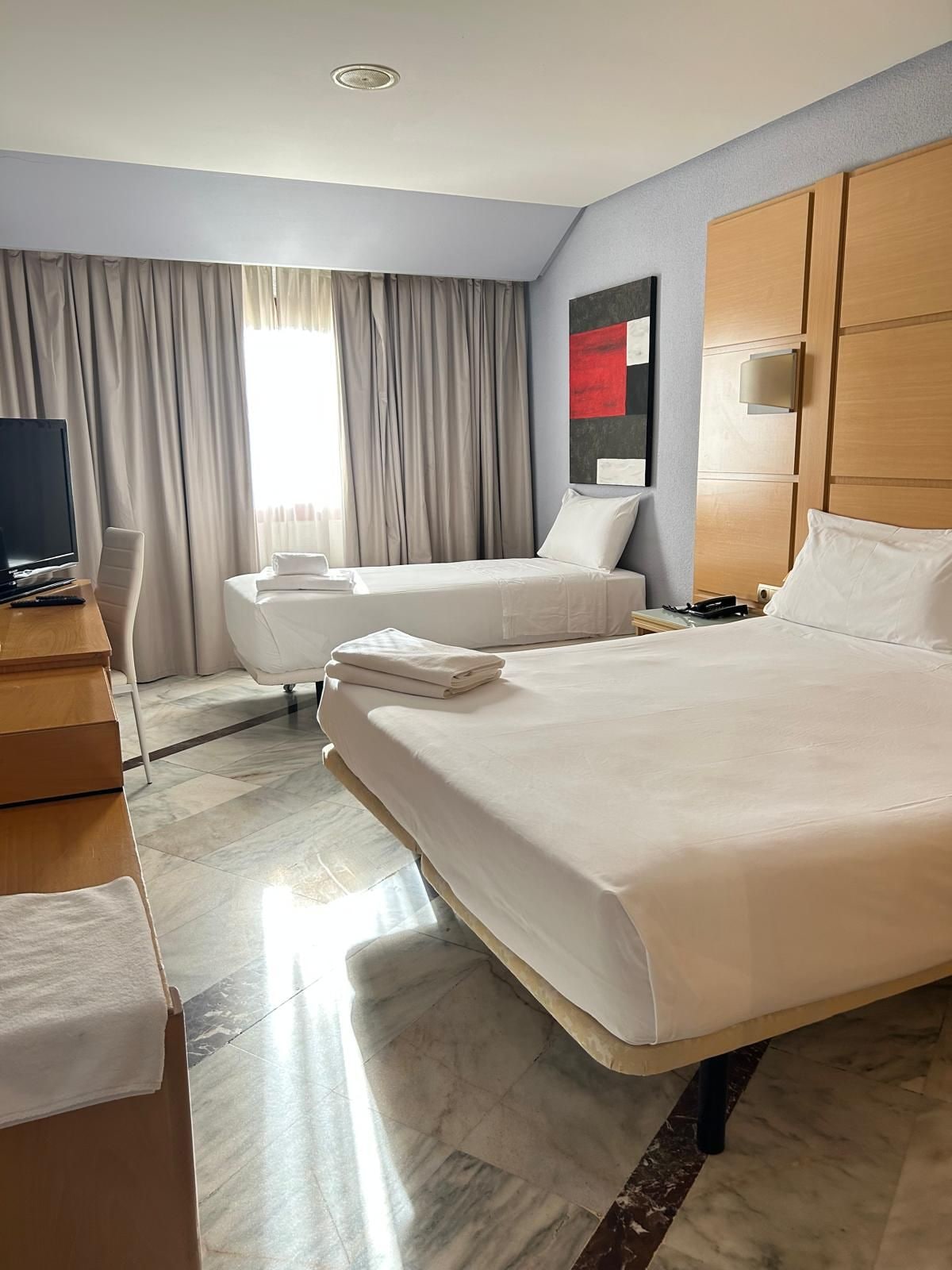 Ein Hotelzimmer mit zwei Betten, einem Schreibtisch, einem Fernseher und einem Fenster mit Vorhängen.