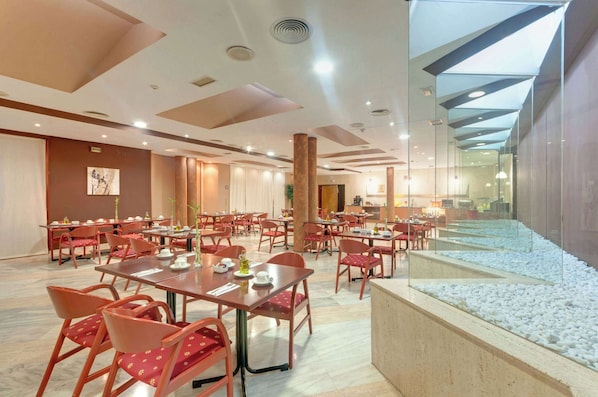 A spacious dining area with multiple tables and chairs, a large glass wall, and a pebbled walkway. - Hotel Puertollano (Puertollano)