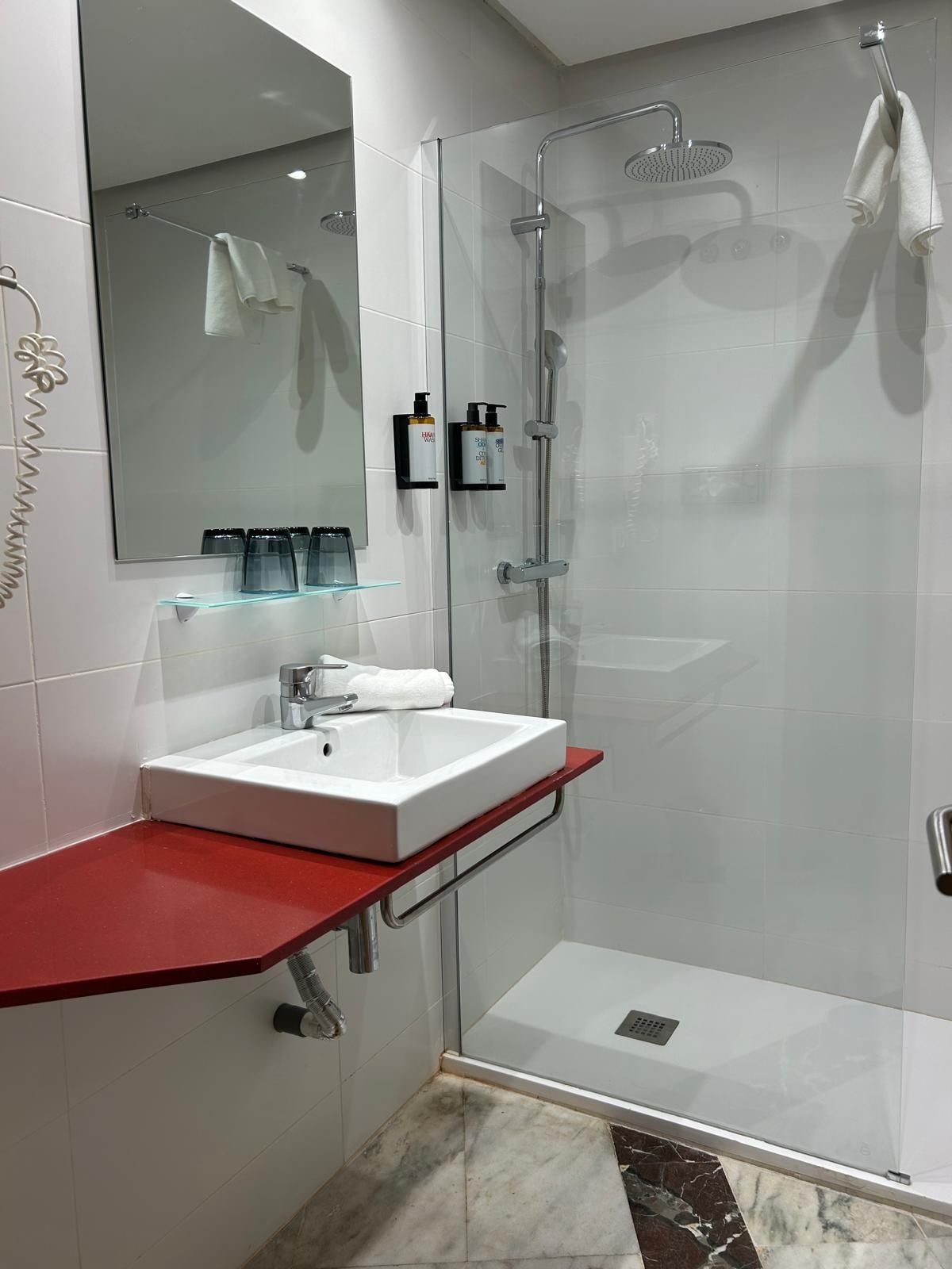 Un baño moderno con ducha de vidrio, un lavabo blanco con encimera roja y un espejo sobre el lavabo.