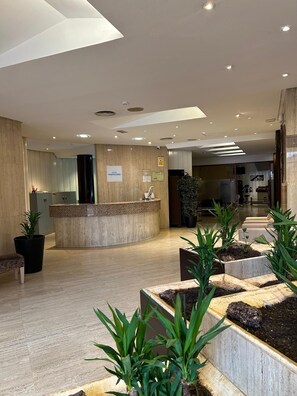 A hotel reception area with a curved counter, potted plants, and a sign indicating the hotel's name. - Hotel Puertollano 3* by eme hoteles (Puertollano)