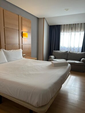 A hotel room with a large bed, a sofa, a nightstand, and a lamp. - Hotel Puertollano 3* by eme hoteles (Puertollano)