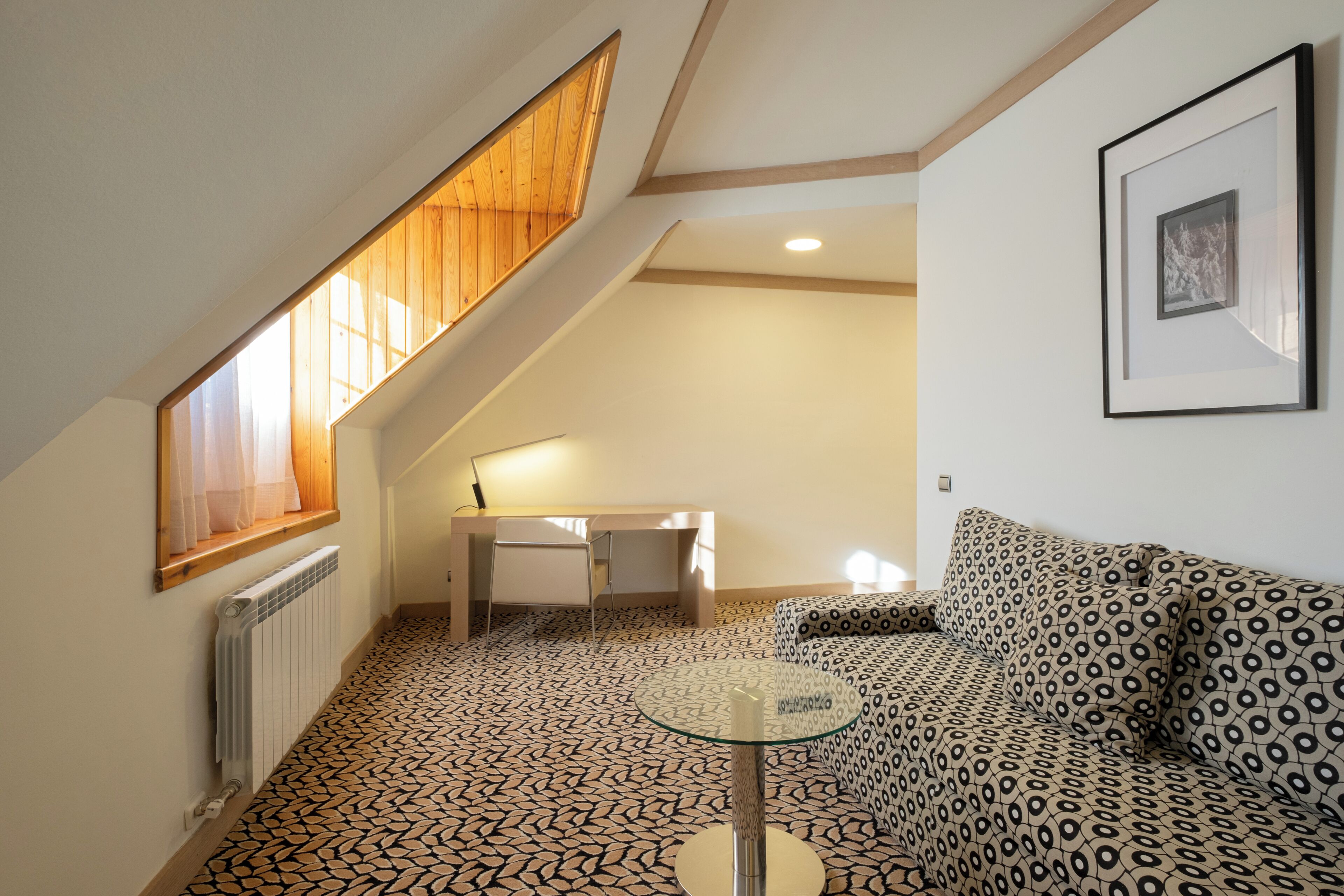 A room with a sloped ceiling, a patterned floor, a glass table, a sofa, a desk, and a framed picture on the wall.