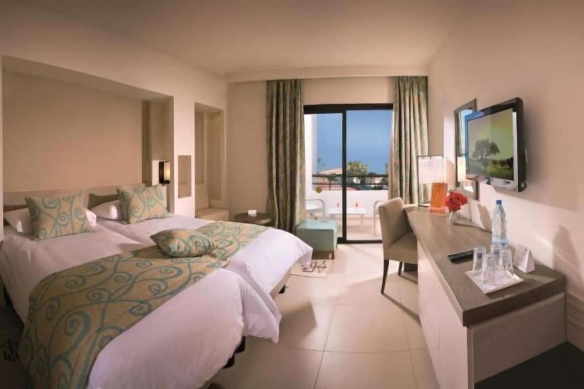 A hotel room with two beds, a desk, a chair, a TV, and a balcony with a view.
