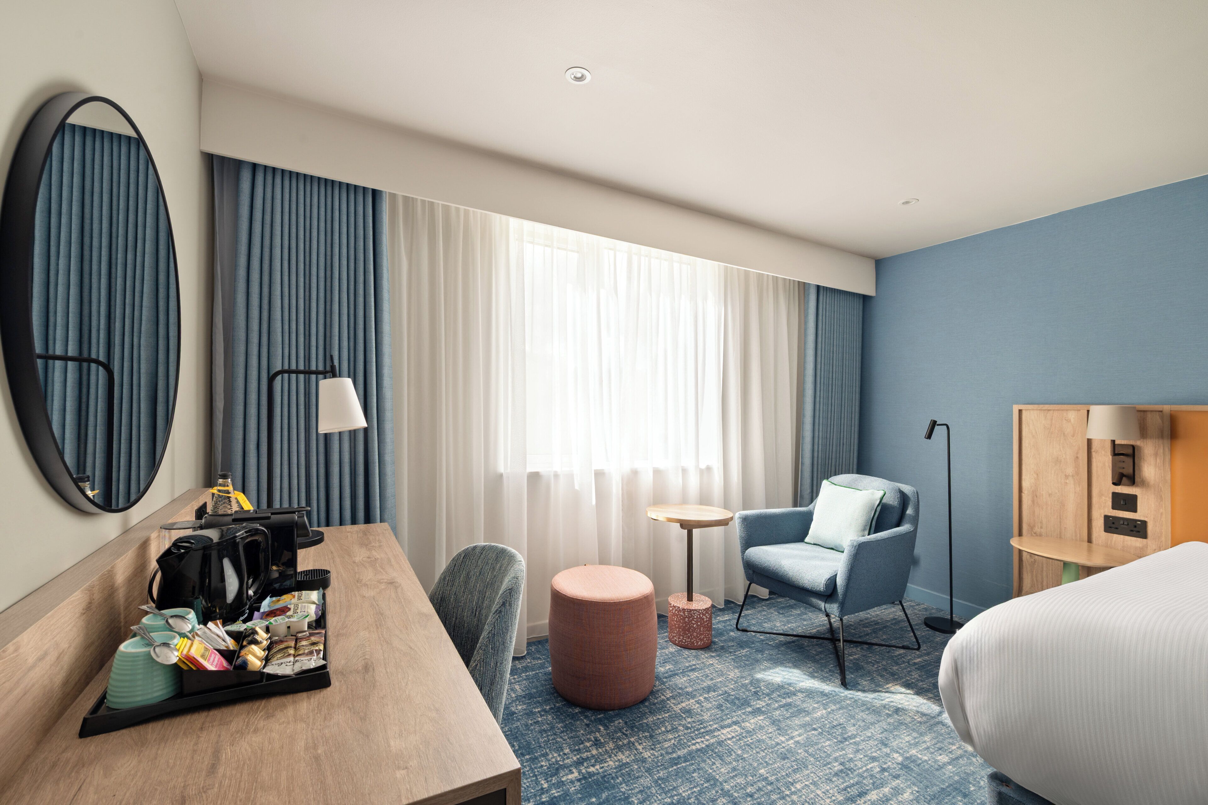 A hotel room with a bed, a desk with a coffee maker, a chair, a round ottoman, a small table, and a mirror.