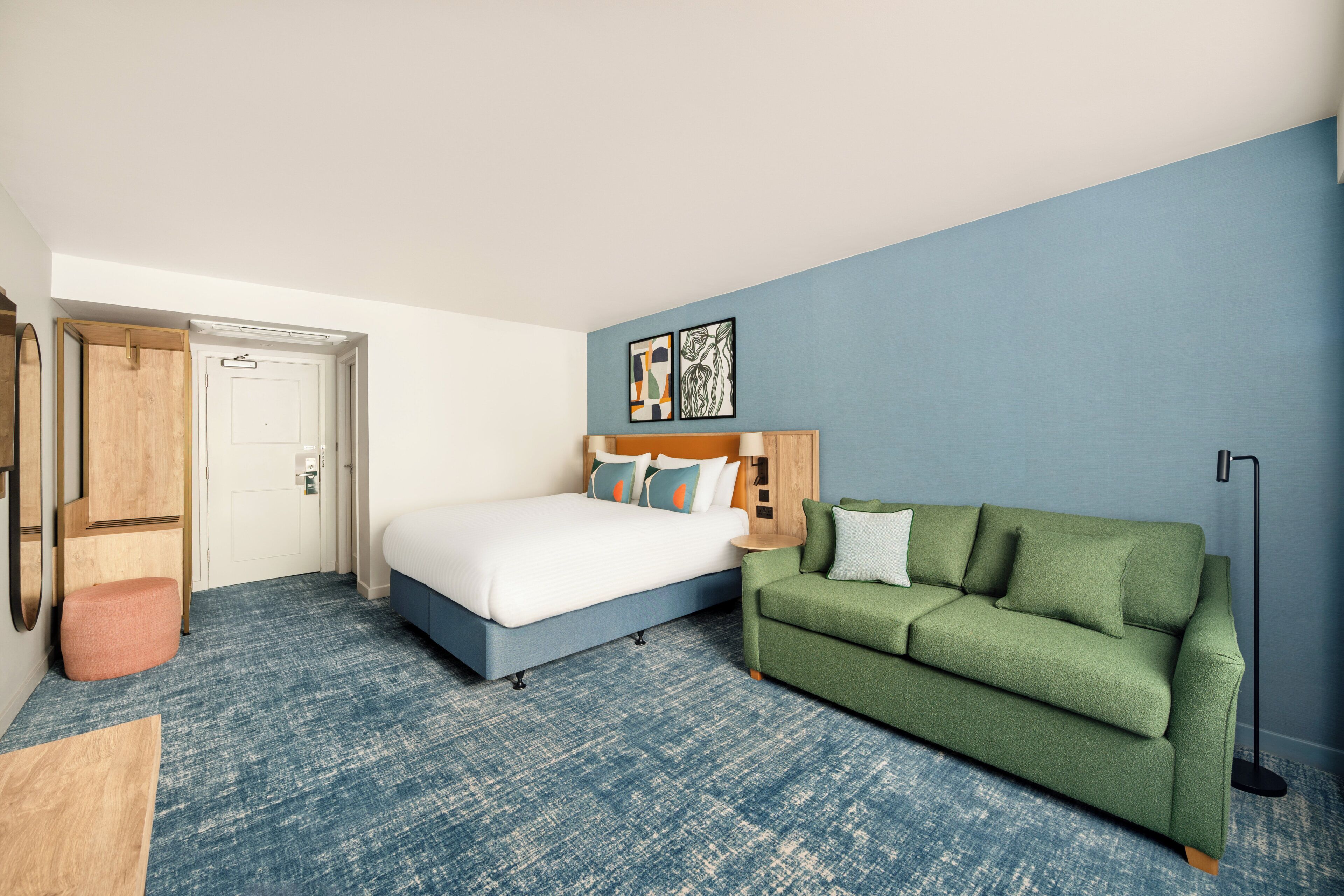 Una habitación de hotel con una cama, un sofá verde, una puerta de madera y una alfombra azul con textura.
