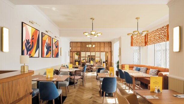 A modern dining area with wooden tables, blue chairs, and a central seating area with orange cushions.