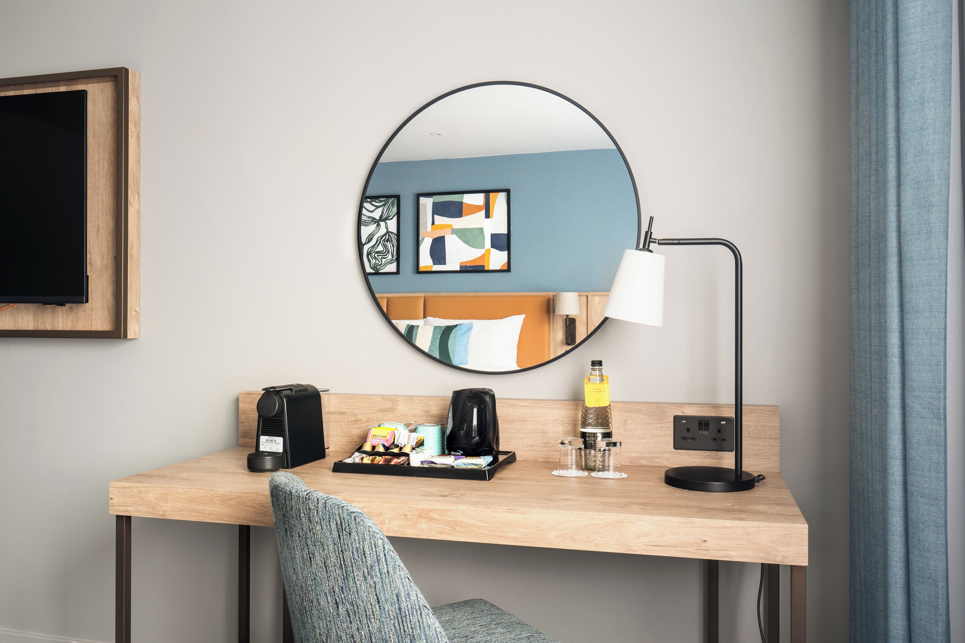 A hotel room with a wooden desk, a round mirror, a coffee maker, a lamp, and a view of the sea through the window.