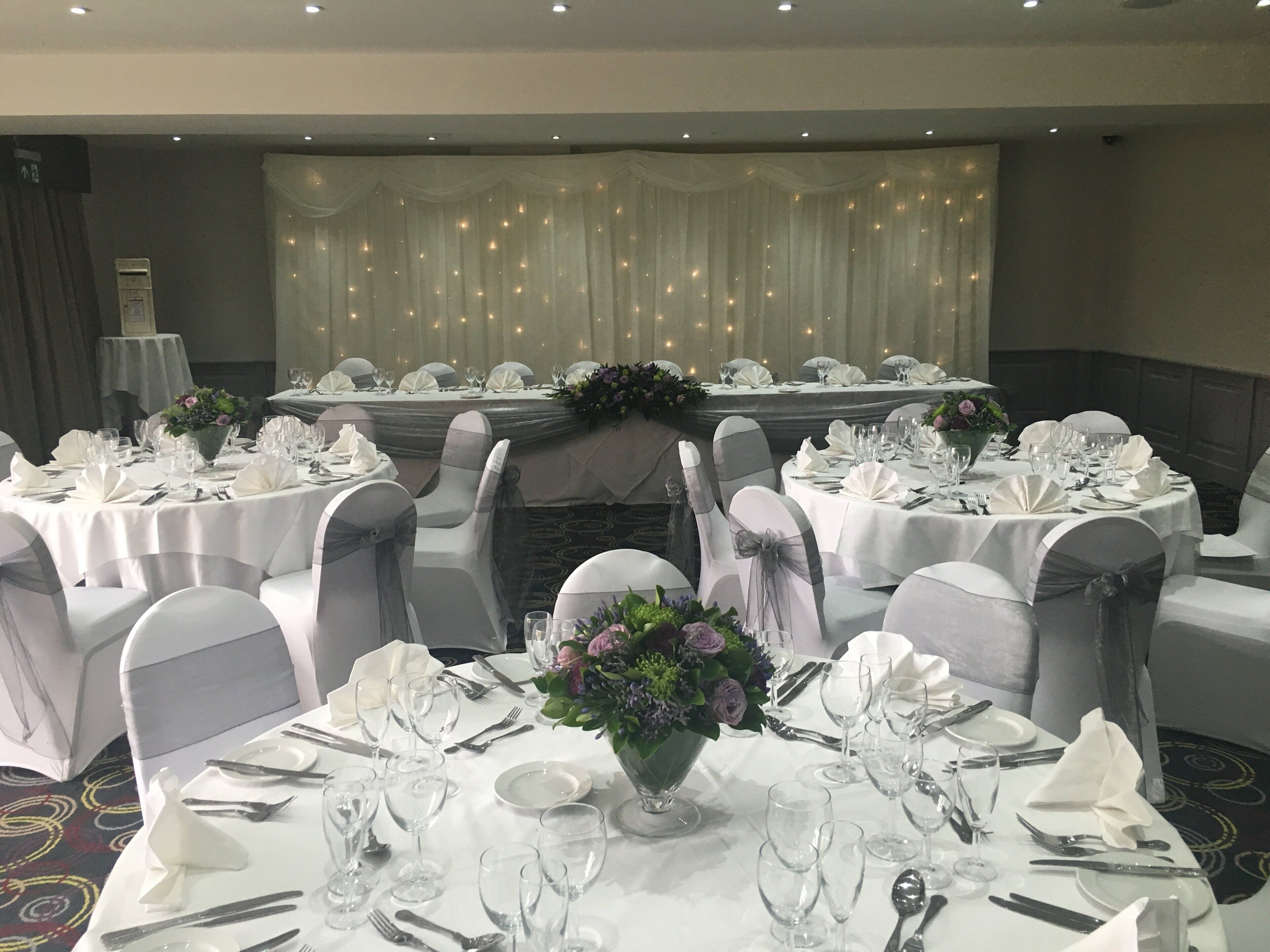 Un salón de banquetes con mesas redondas dispuestas para un evento formal, con manteles blancos, sillas adornadas con cintas grises y centros de mesa florales.