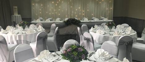 Un salón de banquetes con mesas redondas dispuestas para un evento formal, con manteles blancos, sillas adornadas con cintas grises y centros de mesa florales.