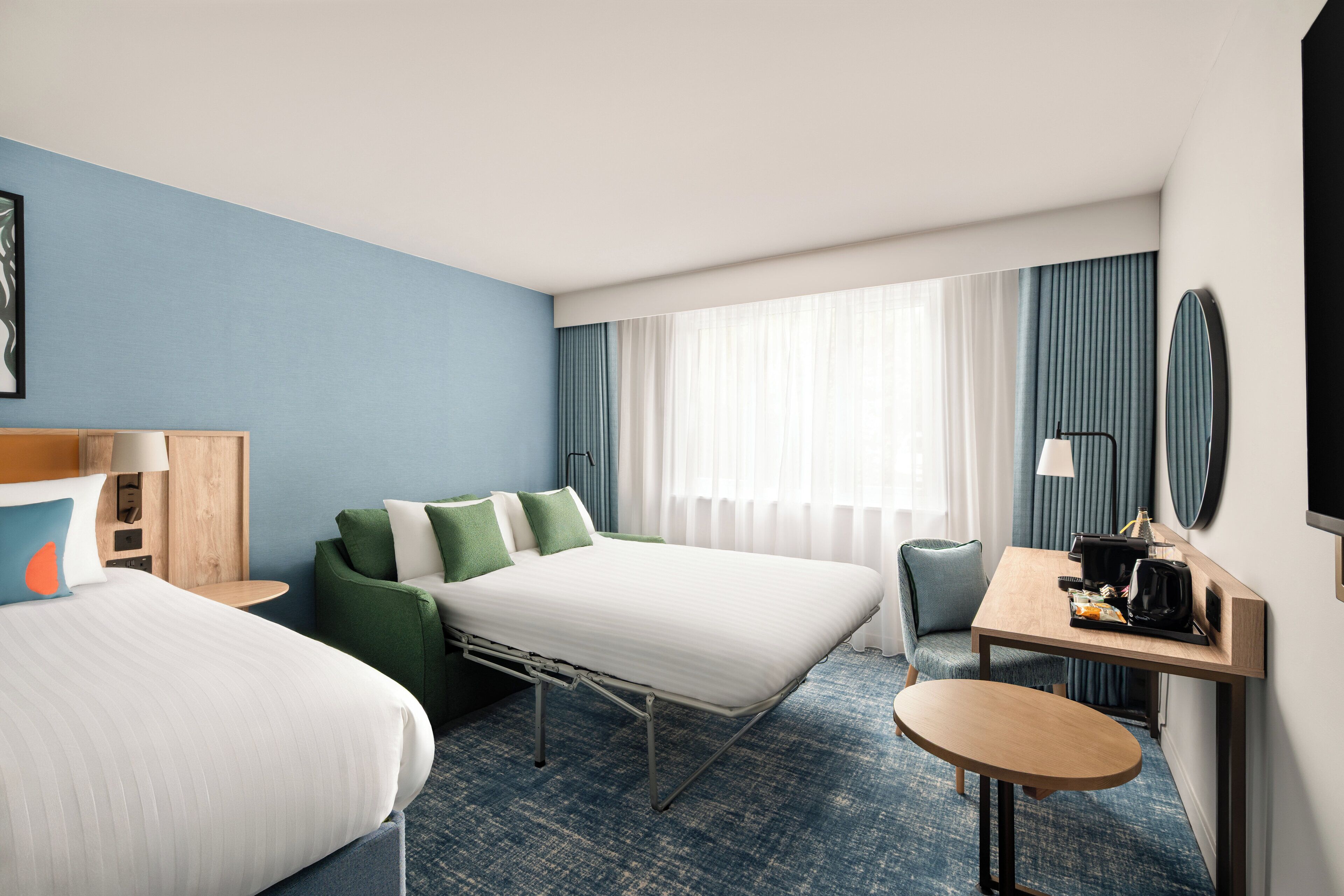 A hotel room with a bed, a desk with a chair, a small table, and a large mirror.